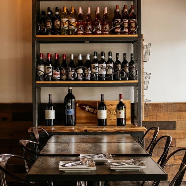 A restaurant interior with a wine display shelf, wooden tables set with cutlery, and metal chairs.